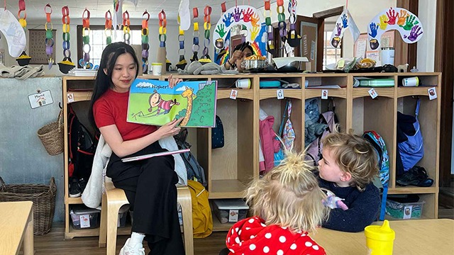 Mavie Pham holds up a children’s book in front of two children seated nearby.