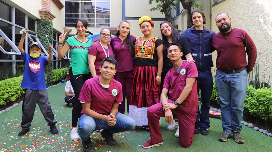A group of people pose for a photo in an outdoor courtyard. Several are wearing maroon scrubs. A child stands nearby on top of confetti on the ground.