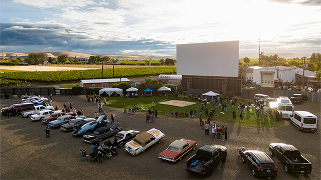 A line of cars parked in front of a large white screen, with vineyards and fields visible in the background.
