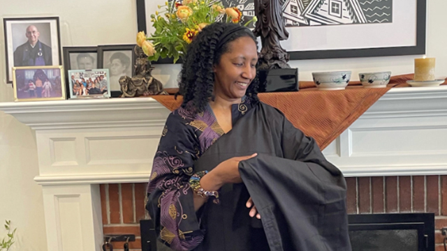 seigen johnson wearing a black robe over purple and black clothes, standing in front of a fireplace and mantle.