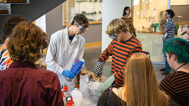 Two students wearing gloves and goggles make ice cream using liquid nitrogen, while other students look on.