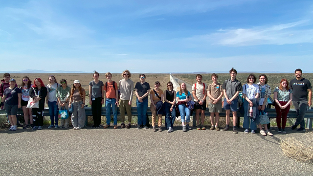 A large group of people stand in front of a fence. A long pipe can be seen stretching into the distance behind them.
