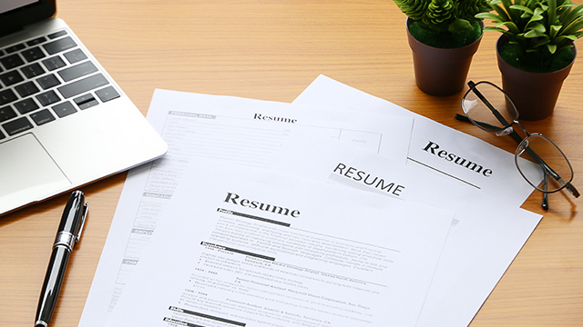 A pile of resumes, a pen, a laptop, glasses and two small potted plants.