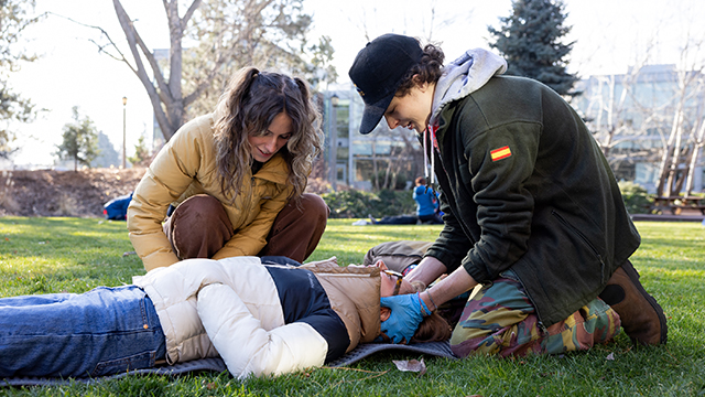 Two students assist another student who is lying on the ground outdoors, in a Wilderness Medicine practice exercise. 