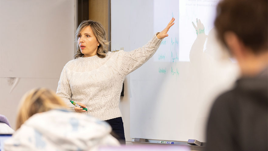 Marina Ptukhina gestures at a whiteboard behind her in a classroom setting.