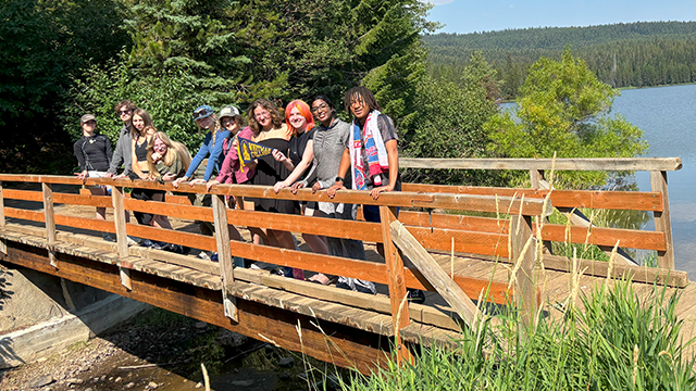 Ten students stand on a wooden pedestrian bridge with trees and a lake in the background.