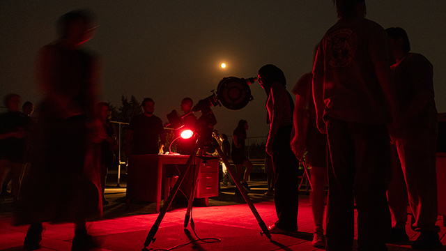 Dimly lit by a red light, students gather around a telescope with the moon visible in the sky behind them. 