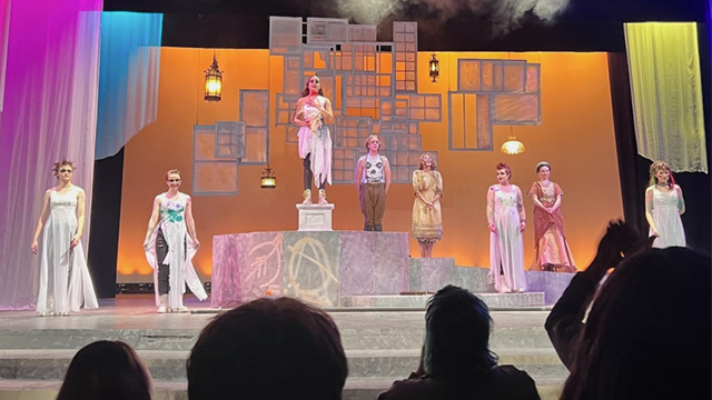 Students stand on stage in Greek-inspired costumes with the silhouettes of applauding audience members in the foreground. 