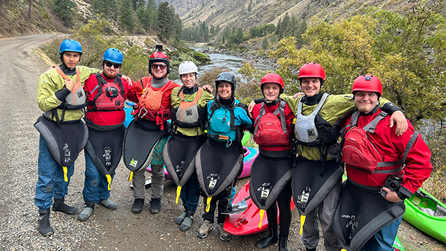 A group of kayakers in protective gear pose for a photo next to a river.