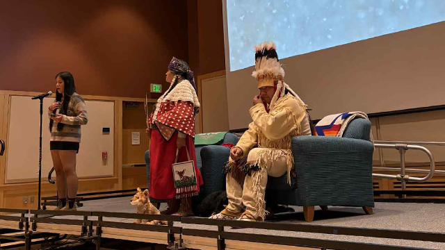 Tatiwyat Buck stands at a microphone on stage with two other figures who are dressed in Native American regalia.