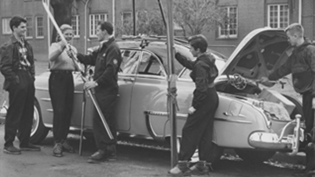 A black and white photo of a group of students with ski equipment loading a vintage automobile.