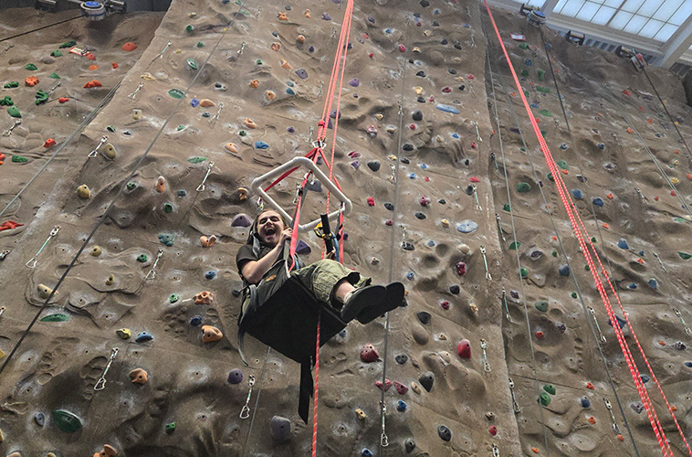 A smiling student in an adaptive harness on a large climbing wall