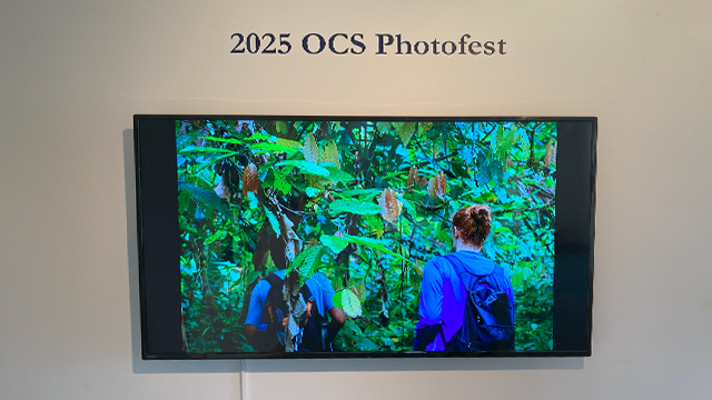 A video monitor displays an image of people walking through a jungle environment. Above it on the wall is the text “2025 OCS Photofest.”