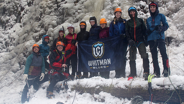 Eleven climbers in protective gear stand in front of a rock wall covered in ice.