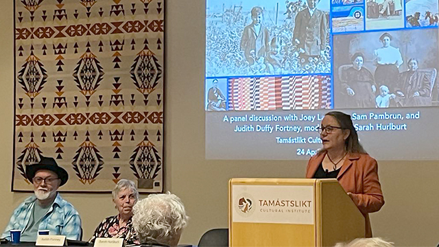 Sarah Hurlburt (right) stands at a lectern giving a presentation with black and white photographs displayed on a screen behind her.