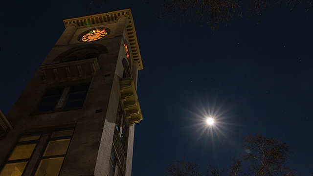 A nighttime image of the Memorial Building clocktower on the left and a full moon (with lens flare) on the right.