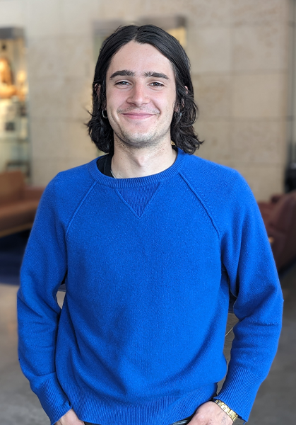 Portrait of Adam Simon with shoulder-length dark hair and wearing a blue sweater.