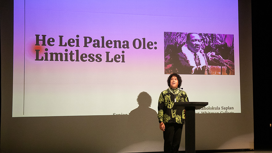 Jace Saplan stands behind a lectern. A slide projected in the background includes an image of Dr. Martin Luther King Jr. wearing a lei and the text “He Lei Palena Ole: Limitless Lei”