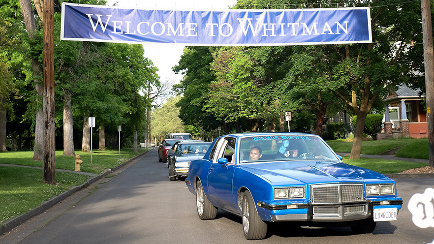 A line of cars driving down a tree-lined street with a large “Welcome to Whitman” banner hanging overhead.