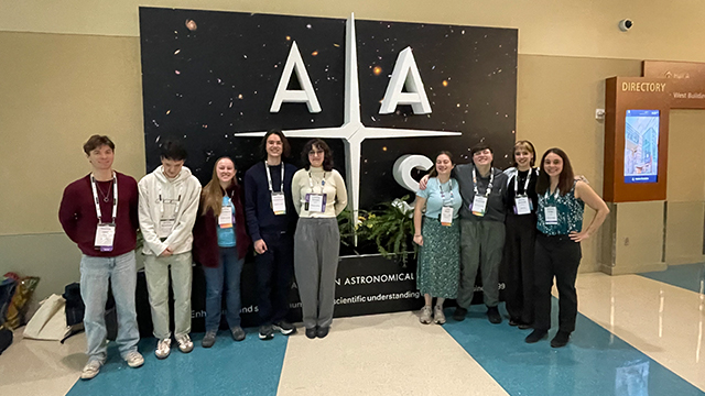 A group of students and their professor stand in front of a black and white display featuring “AAS” large letters
