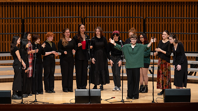 A group of singers, most dressed in black, on a stage standing in front of multiple microphones.