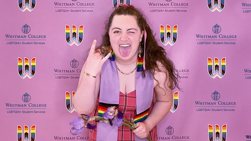 Zoe Perkins ’25 strikes an enthusiastic pose with a lavender and rainbow stole around her neck, standing in front of a lavender backdrop with rainbows and Whitman College logos.