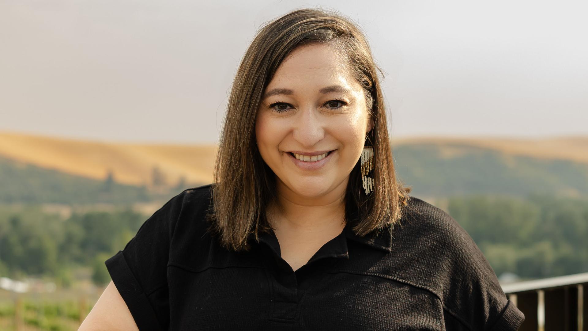 Photo of Abby Muro smiling at the camera and wearing a black shirt dress. There are wheat fields and trees in the background.