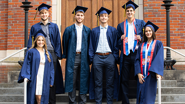 Six students in graduation regalia pose for a photo on the steps in front of a brick building.