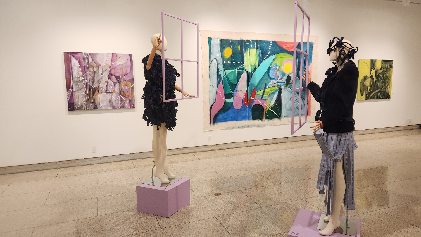 A gallery space with two fashionably-dressed mannequins holding lavender window frames in the foreground and three paintings visible on the wall.