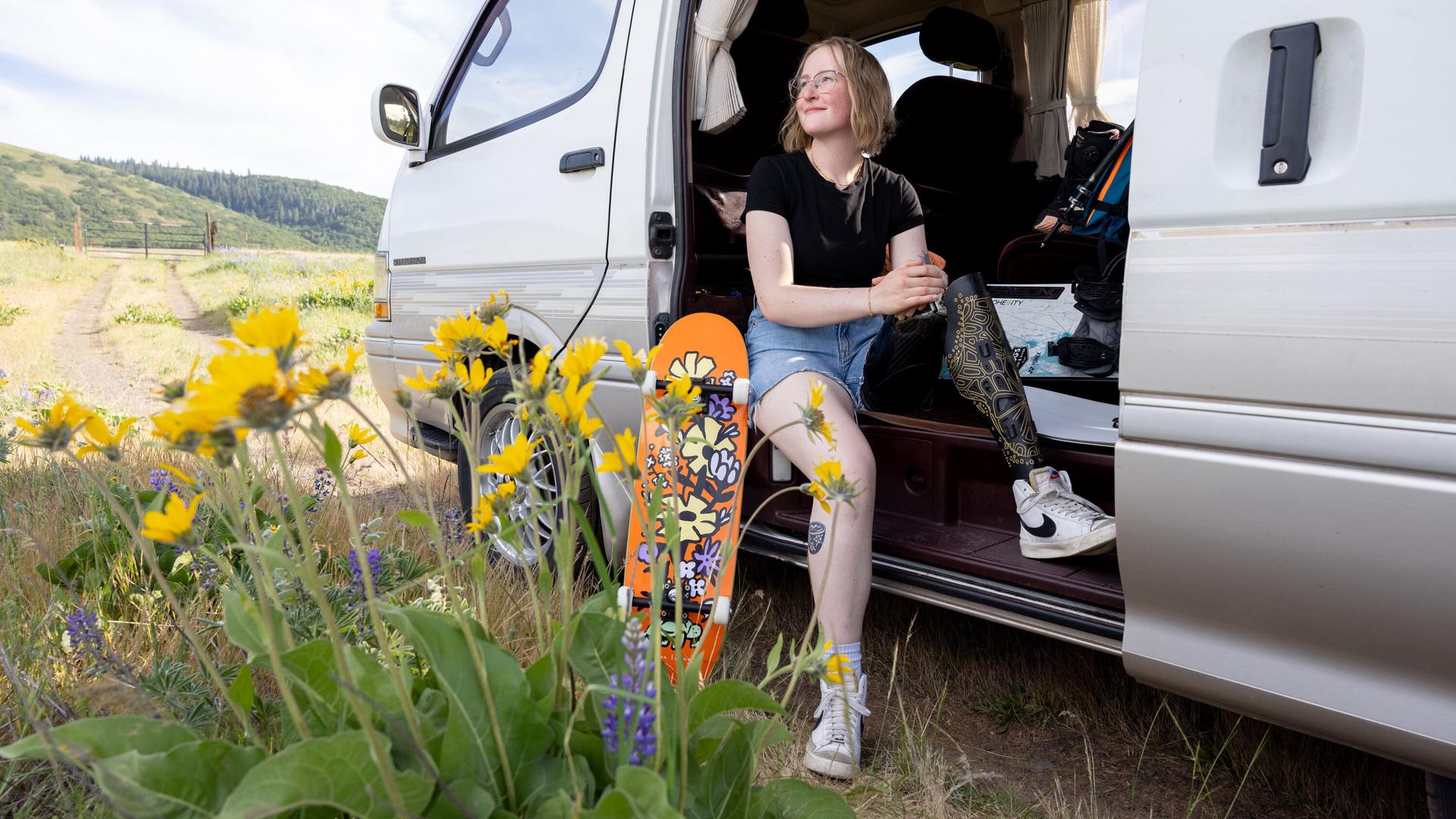 Rebecca Johnston sits in the open doorway of a van parked in an outdoor location with yellow flowers in the foreground.
