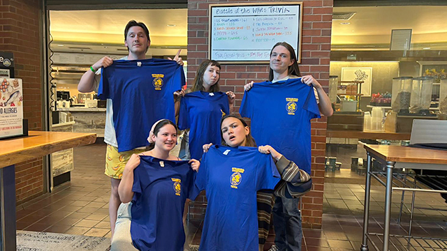 Five students stand in front of a scoreboard, each holding a matching blue t-shirt.