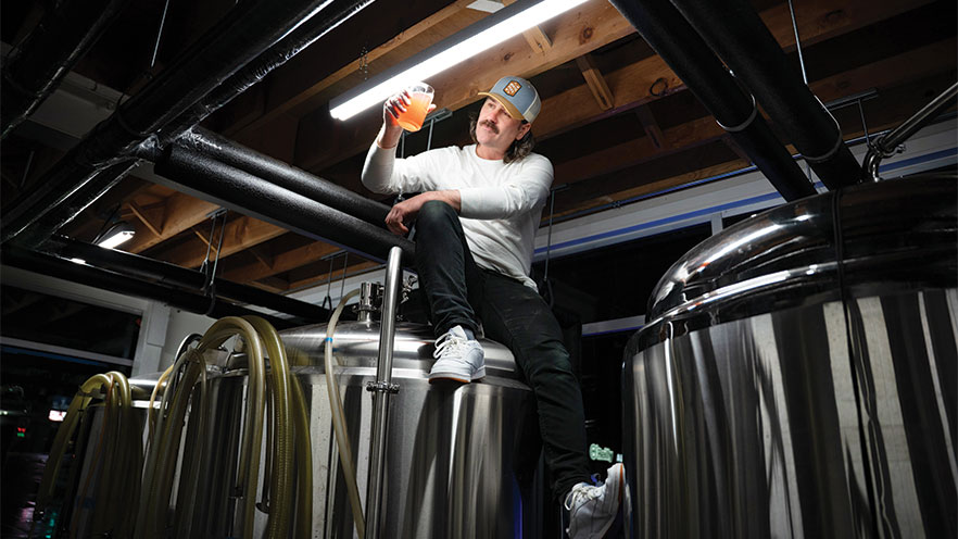 Zachary Lough sits on top of a giant stainless steel container, looking at a glass of cider under a fluorescent light.