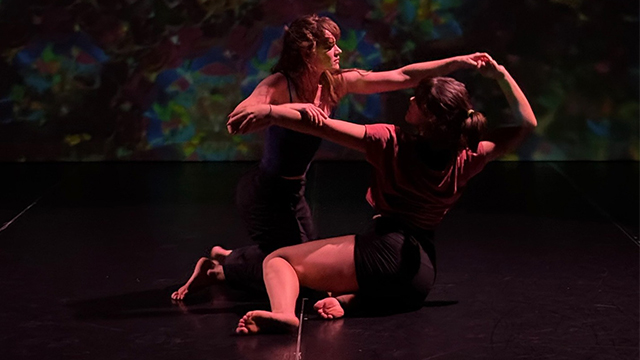 Two dancers in pink light grab each others arms.