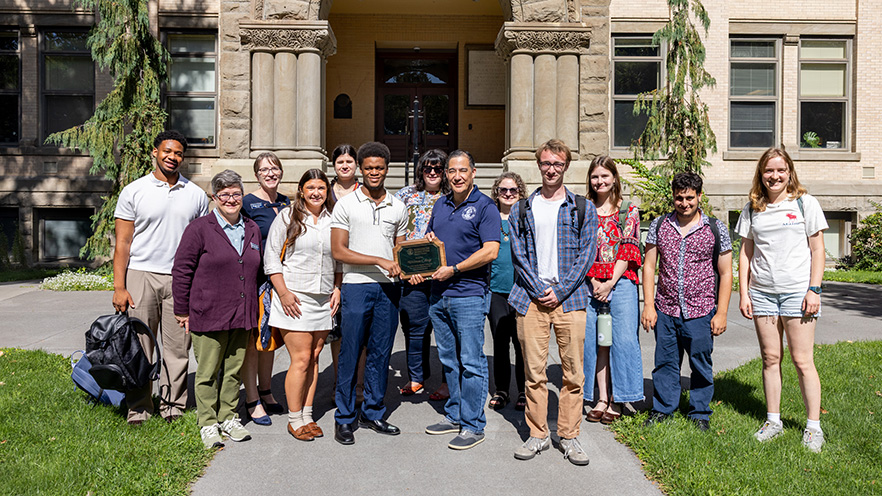A group of people stand in front of the steps of Memorial Hall. In the center, Washington Secretary of State Steve Hobbs hands a plaque to Vinson Russel ’28.