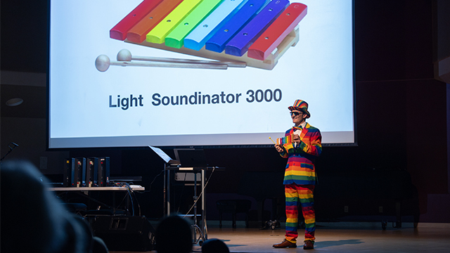 Walker Smith dressed in a rainbow striped outfit stands on a stage with the image of a rainbow-colored xylophone and the words “Light Soundinator 3000” projected on a screen behind them.