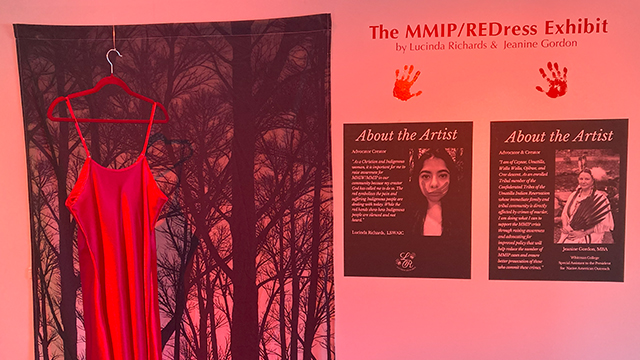 A red-tinted image with a red dress hanging in front of a backdrop of trees on the left and information about the exhibit and artists on the right.