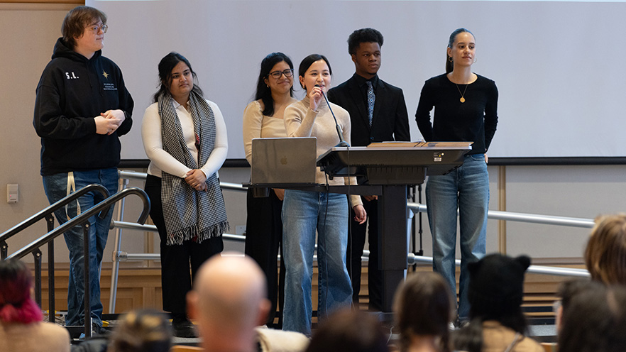 A group of students on stage, one speaks into a microphone.