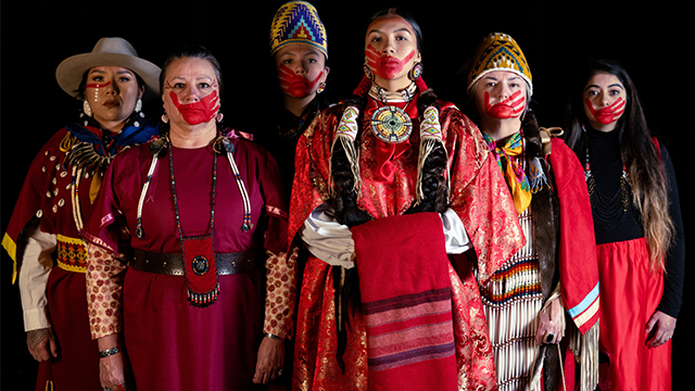 A group of Indigenous women wearing red, most with a red handprint painted on their faces.