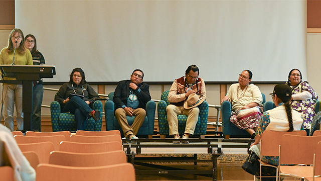 Two students stand at a lectern and five people sit in chairs nearby on stage in the Reid Ballroom.