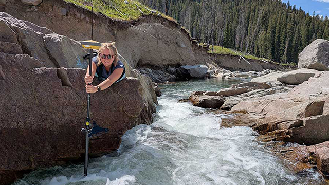 Elly Rectenwald ’26 leans out from a large boulder sticking a pole into the nearby river.