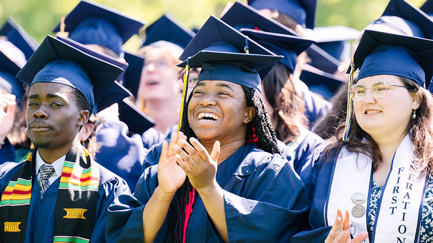 Three students stand in a crowd of other students in graduation regalia.