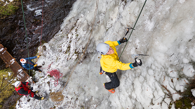 A student climbs up a wall of rough ice, with two people visible on the ground beneath them.