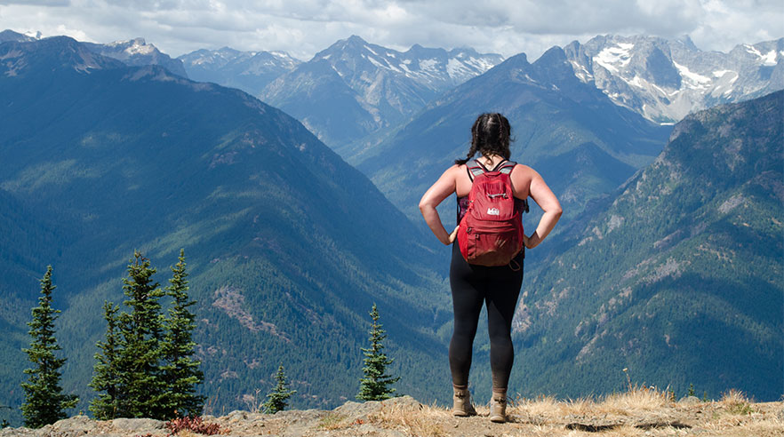 A hiker wearing a backpack stands on a bluff looking at a mountain range.