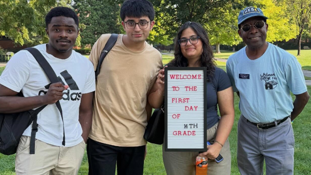 Three students stand with Dean Kazi Joshua (right); one student holds a sign that reads “Welcome to the first day of 14th grade.”