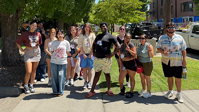 A group of students stand together on a tree-lined sidewalk.