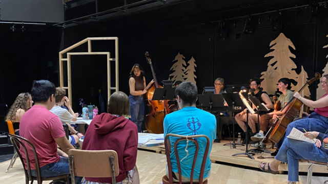 A group of actors sit in chairs near a small orchestra.