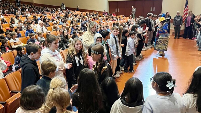 A large group of elementary school children in an auditorium, with two figures up front dressed in Native American regalia.