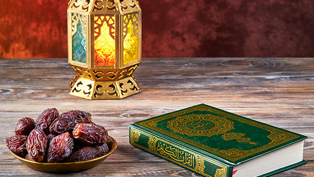 A bowl of dates, a lantern and a copy of the Quran placed on a wooden table.