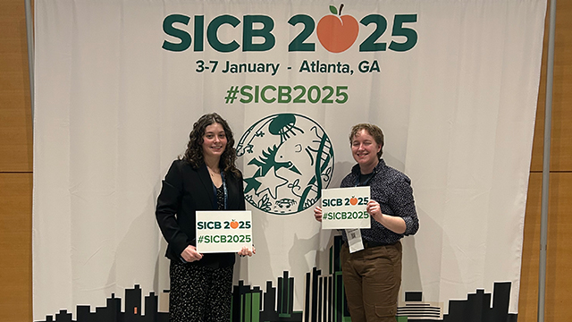 Two students stand in front of a “SICB 2025” banner.