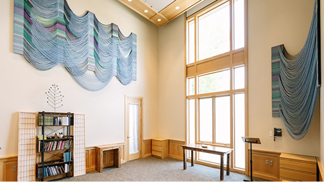 The brightly-lit interior of the All Faiths Room with a large window, a bookcase, and large hanging art pieces on the walls.